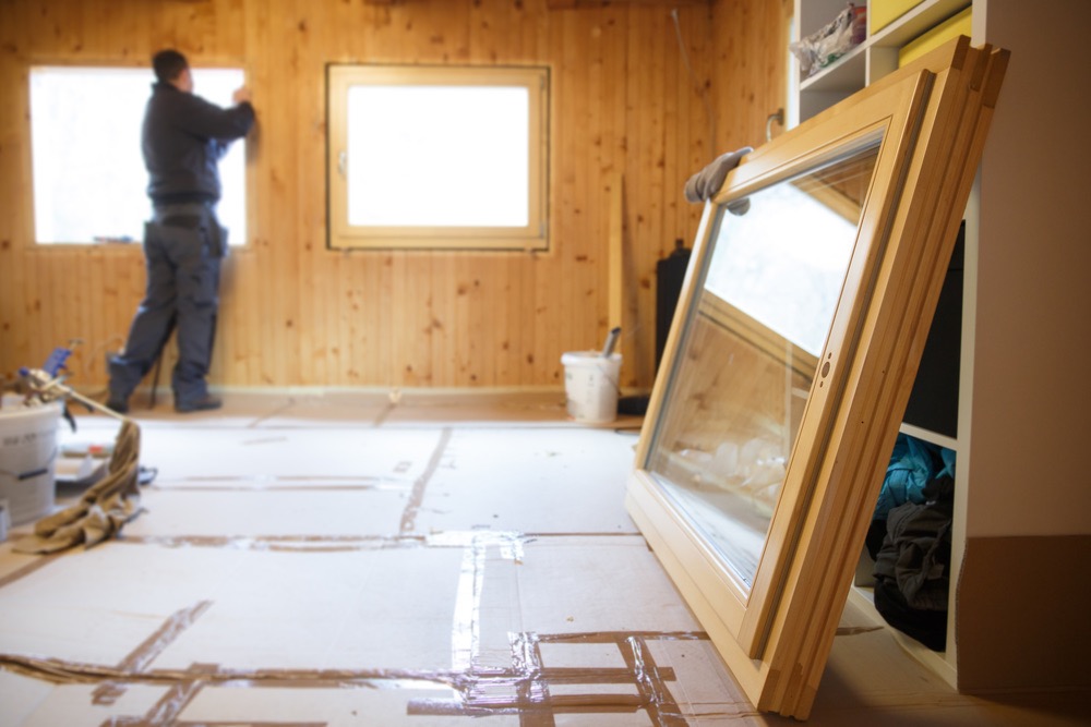Worker in the background installing new, three pane wooden windows in an old wooden house, with a new window in the foreground. Home renovation, sustainable living, energy efficiency concept.